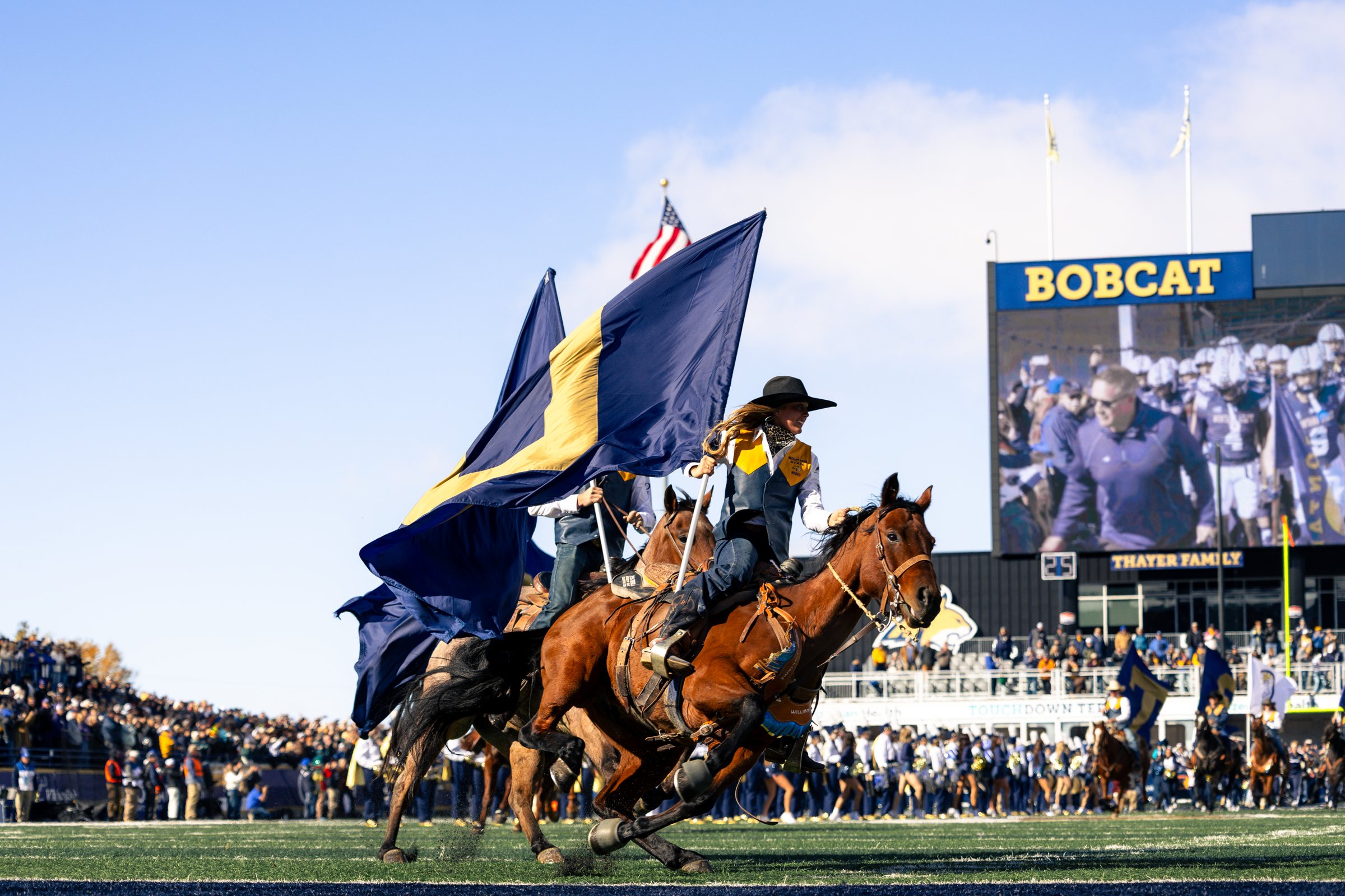 A Bobcat rider carries the flag past the video board